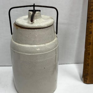 May include: A vintage stoneware jar with a lid and a wire handle. The jar is a light gray color with a crackled surface. The lid is off-white. A wooden ruler is visible in the background.