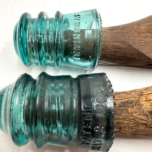 May include: Two antique glass insulators in shades of teal and dark teal, mounted on wooden bases. One insulator is marked "HEMINGRAY", the other "PATENT MAY 1883". These vintage pieces are likely from a telegraph or telephone system.