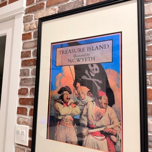 May include: Framed print of "Treasure Island" illustrated by N.C. Wyeth. The artwork depicts pirates with a skull and crossbones flag, set against a blue sky. The print is in a black frame with a white mat, hanging on a brick wall.