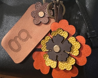 May include: A leather flower keychain with orange, yellow, and brown petals, attached to a black handbag. The keychain includes a small brown flower and a tan tag with the initials "oa" embossed on it.