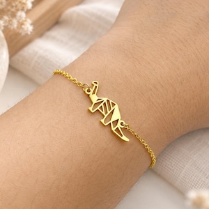 May include: A gold-toned bracelet featuring a delicate chain and a central geometric dinosaur charm. The charm is designed with clean, angular lines that form the silhouette of a long-necked dinosaur, connected to the chain on both sides.