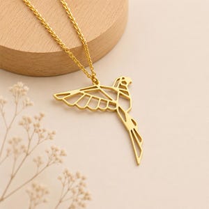 K&ouml;nnte beinhalten: Eine goldfarbene Halskette mit einem Anh&auml;nger in Form eines fliegenden Vogels, der mit geometrischen Linien und offenen Bereichen gestaltet ist. Der Anh&auml;nger ist an einer zarten goldfarbenen Kette befestigt und bildet ein stimmiges Schmuckst&uuml;ck.
