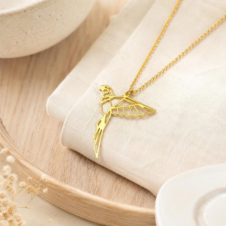 K&ouml;nnte beinhalten: Eine goldfarbene Halskette mit einem zarten, geometrischen Anh&auml;nger in Form eines fliegenden Vogels. Der Anh&auml;nger hat ein offenes, kantiges Design, das an Origami erinnert, und h&auml;ngt an einer d&uuml;nnen, passenden goldfarbenen Gliederkette.