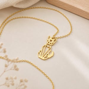 Puede incluir: Un collar de tono dorado con un colgante en forma de gato estilizado. El colgante est&aacute; construido con l&iacute;neas geom&eacute;tricas finas que forman una silueta calada de un felino sentado con orejas puntiagudas y una cola curva unido a una cadena.