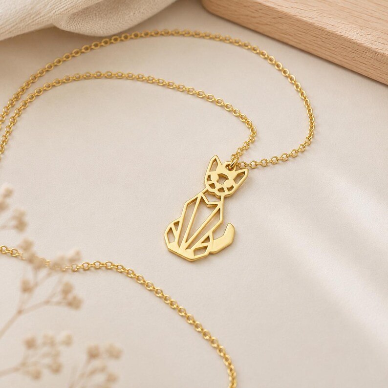 Puede incluir: Un collar de tono dorado con un colgante en forma de gato estilizado. El colgante est&aacute; construido con l&iacute;neas geom&eacute;tricas finas que forman una silueta calada de un felino sentado con orejas puntiagudas y una cola curva unido a una cadena.
