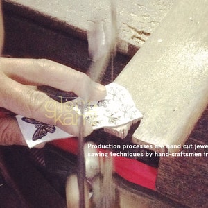 Peut inclure: Gros plan sur une main tenant un morceau de papier avec un motif de papillon. Le papier est coup&eacute; &agrave; la scie. Le texte "Production processes are hand cut jewelry sawing techniques by hand-craftsmen in Thailand." est visible sous le papier.