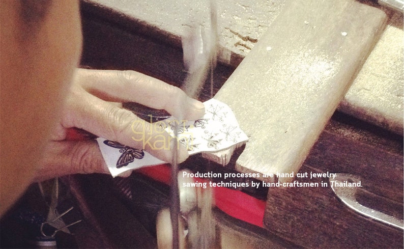Peut inclure: Gros plan sur une main tenant un morceau de papier avec un motif de papillon. Le papier est coup&eacute; &agrave; la scie. Le texte "Production processes are hand cut jewelry sawing techniques by hand-craftsmen in Thailand." est visible sous le papier.