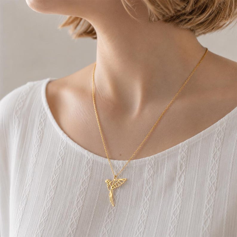 K&ouml;nnte beinhalten: Eine goldfarbene Halskette mit einem zarten, geometrischen Vogelanh&auml;nger in durchbrochenem Design. Der Anh&auml;nger h&auml;ngt an einer d&uuml;nnen, passenden goldfarbenen Kette, die auf dem Schl&uuml;sselbein aufliegt und eine klare, kantige &Auml;sthetik zeigt.