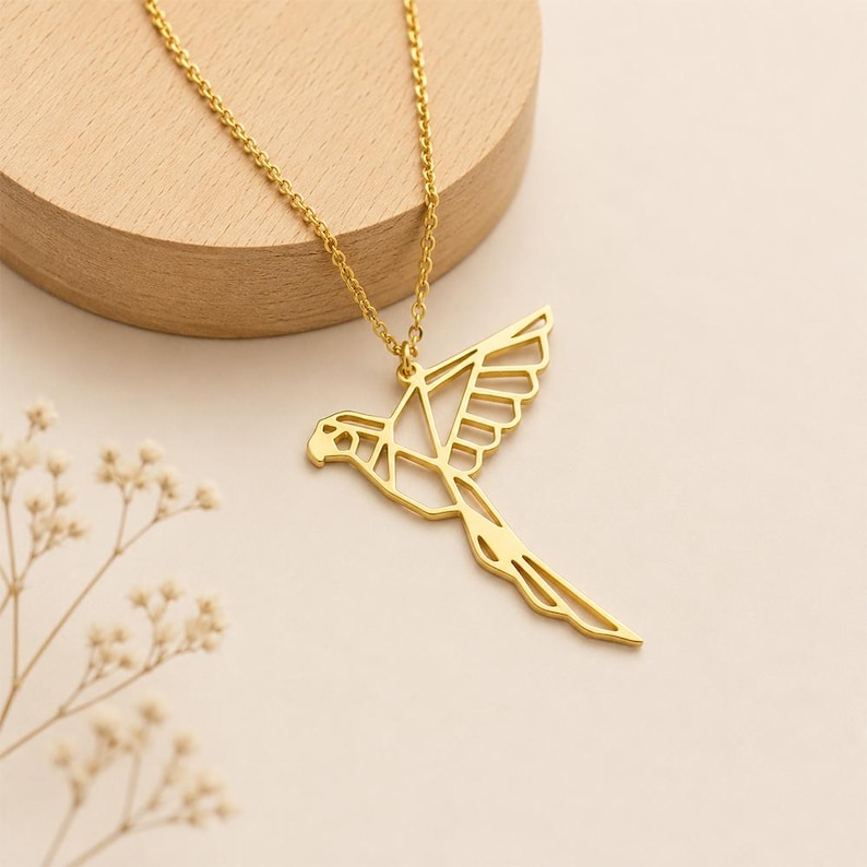 K&ouml;nnte beinhalten: Eine goldfarbene Halskette mit einem geometrischen, durchbrochenen Anh&auml;nger in Form eines fliegenden Vogels. Der Anh&auml;nger h&auml;ngt an einer zarten, passenden goldfarbenen Kette und zeigt klare Linien sowie eine stilisierte, kantige Papagei-Silhouette.