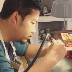 K&ouml;nnte beinhalten: Eine Person in einem blaugr&uuml;nen Langarmshirt und einer beigen Sch&uuml;rze verwendet einen Handbrenner, um an einem Metallst&uuml;ck auf einer Werkbank aus Holz zu arbeiten. Der Arbeitsplatz umfasst eine graue Lampe, verschiedene Werkzeuge und eine rote Klemme.