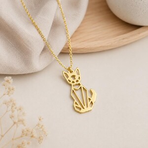 Puede incluir: Un collar de tono dorado con un colgante en forma de gato, dise&ntilde;ado con l&iacute;neas geom&eacute;tricas y espacios abiertos. El colgante est&aacute; unido a una delicada cadena de tono dorado, creando una apariencia simple y estructurada para esta pieza de joyer&iacute;a.