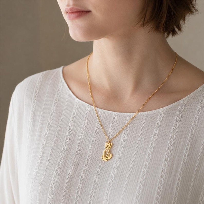 Puede incluir: Un collar de tono dorado con una cadena fina y un peque&ntilde;o colgante plano con forma de gato estilizado de l&iacute;neas geom&eacute;tricas y angulares. El colgante cuelga en el centro de la cadena, que descansa sobre una blusa blanca de manga larga texturizada.