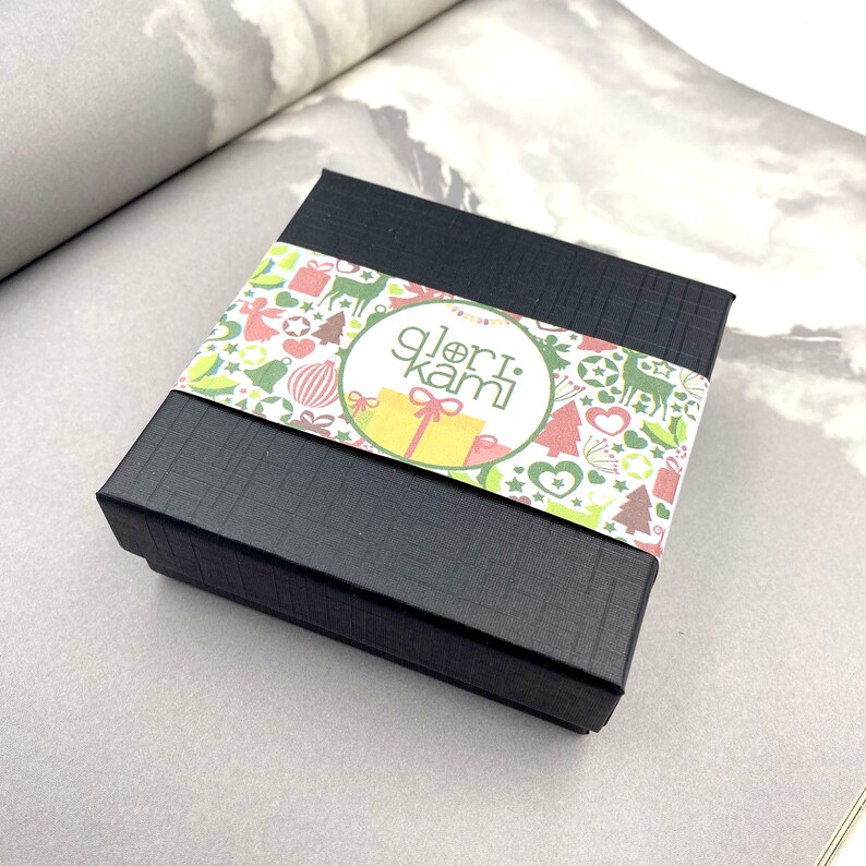 Peut inclure: Une bo&icirc;te cadeau noire avec une &eacute;tiquette &agrave; motifs blancs et verts. L'&eacute;tiquette pr&eacute;sente un cercle avec le texte "glori.kami" en blanc.