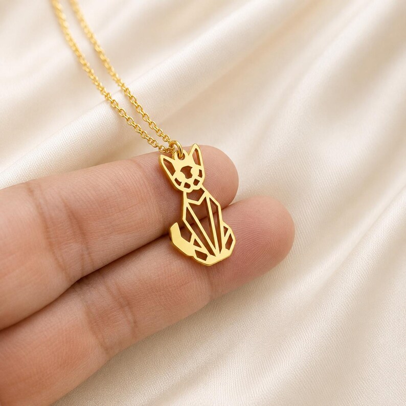 Puede incluir: Un collar de tono dorado con un colgante en forma de gato estilizado con l&iacute;neas geom&eacute;tricas caladas. El colgante cuelga de una delicada cadena de tono dorado, mostrando un dise&ntilde;o limpio y angular que forma la silueta de un gato sentado.