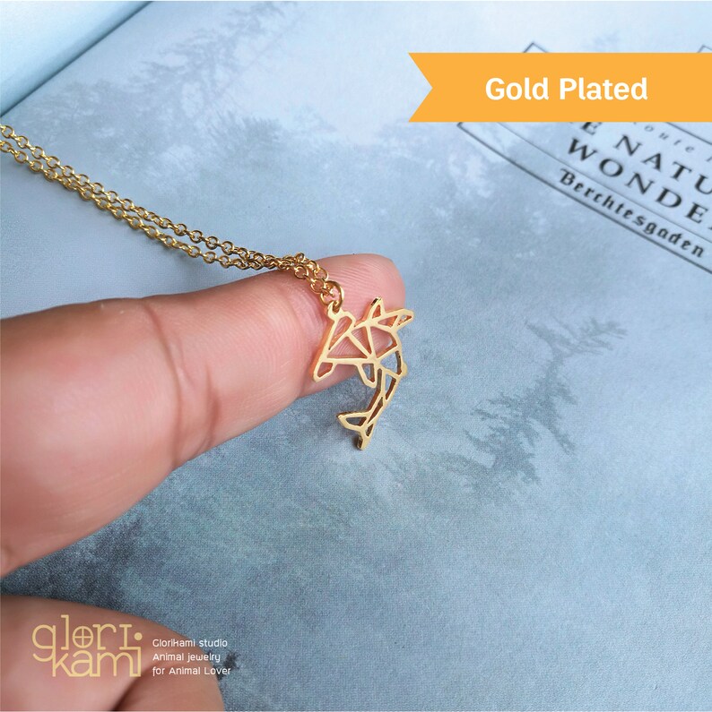 Peut inclure: Collier plaqu&eacute; or avec un pendentif en forme de dauphin g&eacute;om&eacute;trique. Le collier est sur un doigt et l'arri&egrave;re-plan est un livre avec le texte "E NATU WONDE Berchtesgaden".