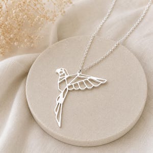 K&ouml;nnte beinhalten: Eine silberfarbene Halskette mit einem Anh&auml;nger in Form eines fliegenden Vogels. Der Anh&auml;nger besteht aus d&uuml;nnen, miteinander verbundenen Metalllinien, die die geometrische Silhouette eines Papageis mit ausgebreitetem Fl&uuml;gel bilden.