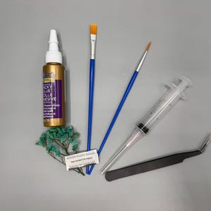 May include: A crafting kit containing a bottle of Aleene's Original Tacky Glue, two blue-handled paintbrushes, a clear plastic syringe, a pair of black angled tweezers, a small rectangular packet labeled The Picketts Press, and a sprig of dried teal flowers.