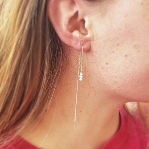 May include: A silver chain earring with three small white pearls hanging from it. The earring is worn in the left ear of a person with long blonde hair.