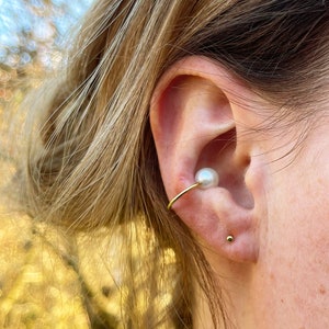 Könnte beinhalten: Ein goldener Ohrring mit einer weißen Perle, die oben befestigt ist. Der Ohrring wird am oberen Teil des Ohrs getragen, und ein kleiner goldener Ohrstecker wird im unteren Ohrläppchen getragen.