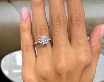 May include: A close-up of a hand wearing a silver engagement ring. The ring features a large, round, clear gemstone as the centerpiece, with smaller stones set along the band. The nails are painted with a light pink polish.