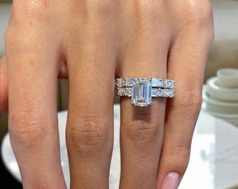 May include: A two-piece diamond ring set. The engagement ring features a large emerald-cut diamond center stone, flanked by smaller diamonds. The matching band is set with multiple smaller diamonds. The rings are set in a silver metal.