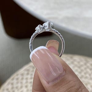 Puede incluir: Un anillo de tono plateado que presenta dos piedras redondas transparentes engastadas una al lado de la otra en la parte superior. La banda es delgada y está decorada con una serie de pequeñas piedras transparentes brillantes incrustadas.