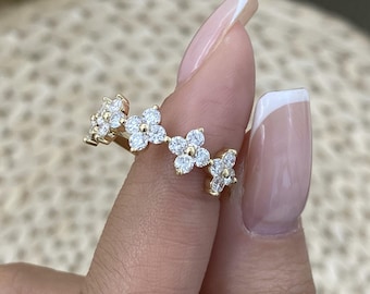 May include: A gold ring featuring a series of four-petaled flower motifs, each constructed from small, clear, round-cut stones. The band is thin and metallic, showcasing a repeating floral pattern that encircles the finger with sparkling accents.