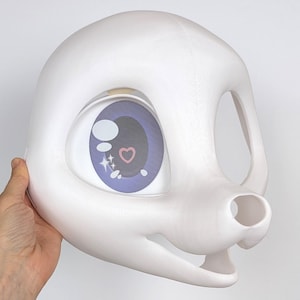 May include: A white, stylized animal mask with a large eye featuring a purple iris, a heart, and star accents. The mask has a smooth, rounded design with open areas for the mouth and the other eye.
