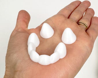 Puede incluir: Un juego de colmillos de vampiro de plástico blanco y dos colmillos, en una palma abierta. Los dientes están diseñados para encajar sobre los dientes superiores, y los colmillos son separados. Para disfraces o uso de novedad.
