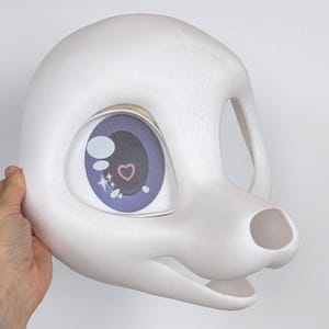 May include: A white, blank fursuit head with a large eye featuring a purple iris and a heart design. The head has a rounded shape with openings for the eyes, mouth, and ears. The eye design includes a heart and star accents.
