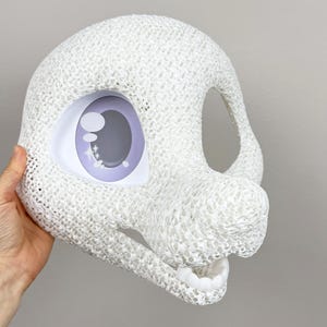Könnte beinhalten: Eine weiße, strukturierte Maske mit einem großen Auge mit einer lila Iris und weißen Highlights. Die Maske hat einen offenen Mund und eine große Öffnung für das andere Auge. Die Maske wird von einer Hand vor einem grauen Hintergrund gehalten.