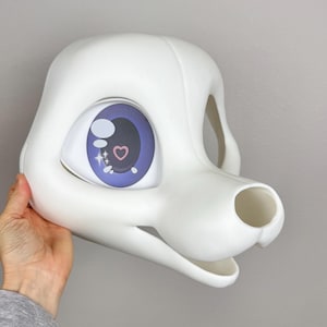 May include: A white, blank fursuit head with large, expressive eyes. The eyes are a light purple with a heart and star design. The head has a smooth, clean finish and is designed to fit over the wearer's head.