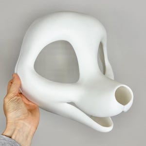 May include: A white, smooth, 3D-printed mask in the shape of a canine head. The mask has large eye openings, a prominent snout, and a detailed jawline. The mask is held by a hand against a plain gray background.