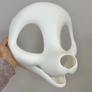 May include: A white, smooth-surfaced mask with large eye openings and a snout-like protrusion. The mask is held by a hand, showcasing its three-dimensional form. The design suggests a stylized animal or creature.