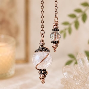 Op de afbeelding: Een koperkleurige metalen ketting hangt aan een grote, bolvormige, gebarsten witte glaskraal omwikkeld met koperdraad, geaccentueerd met donkere metalen kralen. Een kleinere, heldere glaskraal is bevestigd aan een tweede ketting met koperdraad.