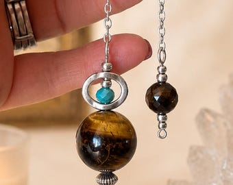 May include: A pendulum featuring a large, spherical tiger's eye stone bead, a small turquoise bead set within a silver-toned metal ring, and a faceted dark brown bead on a silver-toned chain. The design includes decorative silver-toned metal caps.