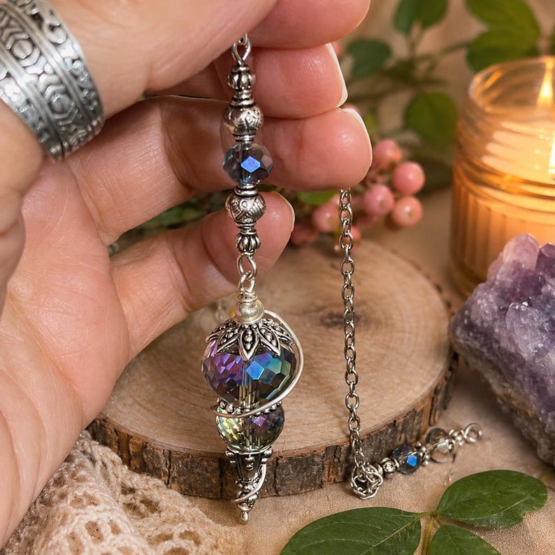 May include: A silver-toned pendulum featuring a faceted, iridescent glass bead at the base, topped with an ornate metal cap. The chain includes smaller dark blue faceted beads and silver metal spacers, creating a decorative hanging ornament.