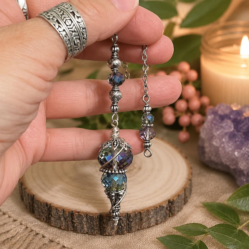 May include: A silver-toned pendulum featuring two faceted, iridescent glass beads in shades of blue and purple, accented with intricate metal filigree and smaller decorative beads on a silver chain, suspended vertically with a metallic finish.