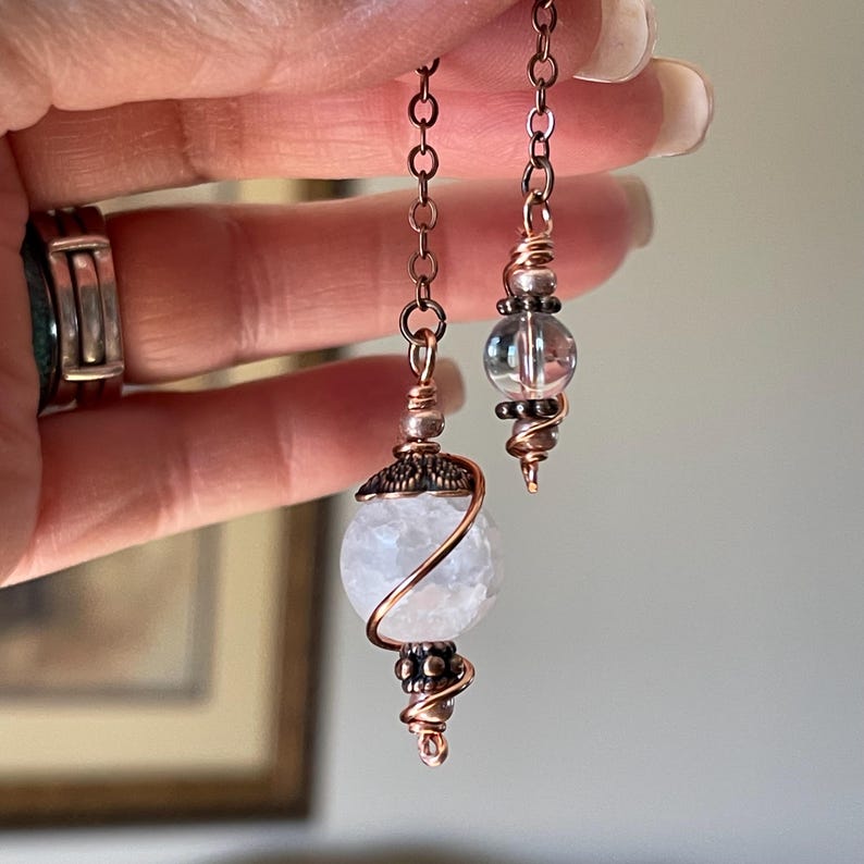Op de afbeelding: Een paar hangende oorbellen met koperdraad en kralen. De grotere oorbel heeft een ronde, getextureerde witte steen omhuld door koperdraad met decoratieve kapjes, terwijl de kleinere oorbel een heldere, gladde glazen kraal bevat.