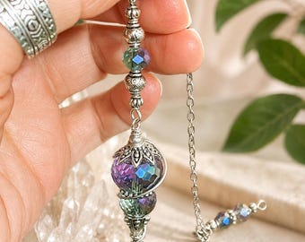 May include: A silver-toned pendulum featuring a faceted, iridescent purple and green glass bead at the base, topped with ornate silver metal caps and smaller matching beads. The pendulum hangs from a silver metal chain with additional small beads.
