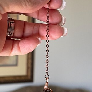 Op de afbeelding: Een pendel met een bolvormige witte craquel&eacute; kwartskraal, omwikkeld met koperdraad en voorzien van decoratieve metalen kapjes. De kraal hangt aan een koperkleurige metalen ketting die aan de bovenzijde eindigt in een kleine doorzichtige kraal.
