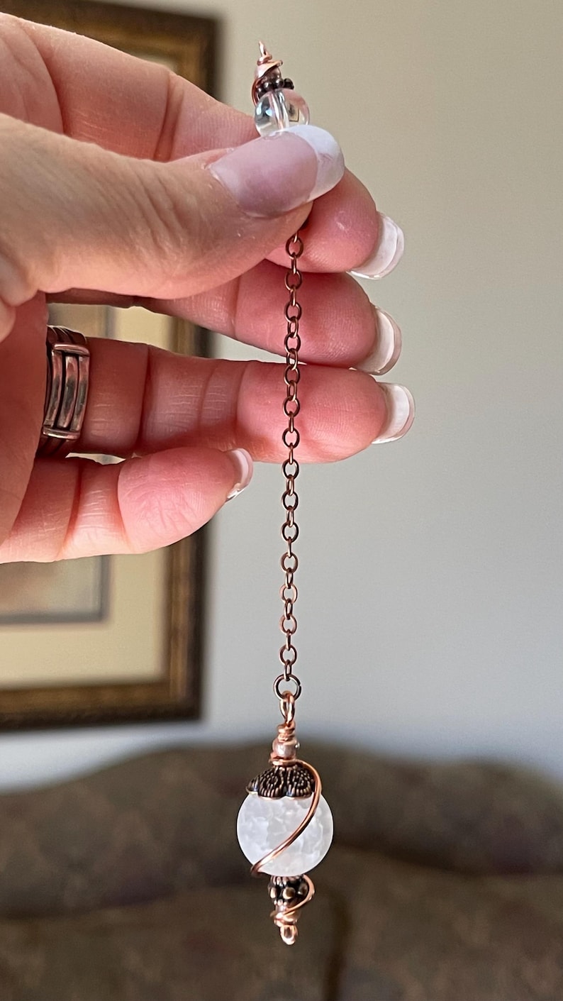 Op de afbeelding: Een pendel met een bolvormige witte craquel&eacute; kwartskraal, omwikkeld met koperdraad en voorzien van decoratieve metalen kapjes. De kraal hangt aan een koperkleurige metalen ketting die aan de bovenzijde eindigt in een kleine doorzichtige kraal.