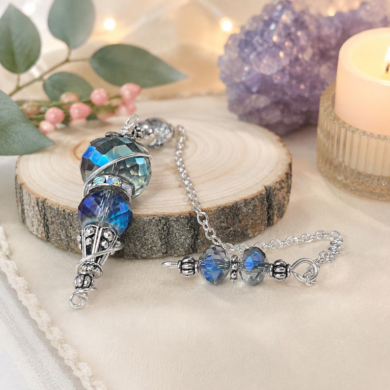 May include: A silver-toned metal pendulum featuring faceted blue and iridescent crystal beads. The main body consists of a large spherical bead and a smaller tapered bead, accented with intricate silver filigree caps. A silver chain includes two additional small blue crystal beads.