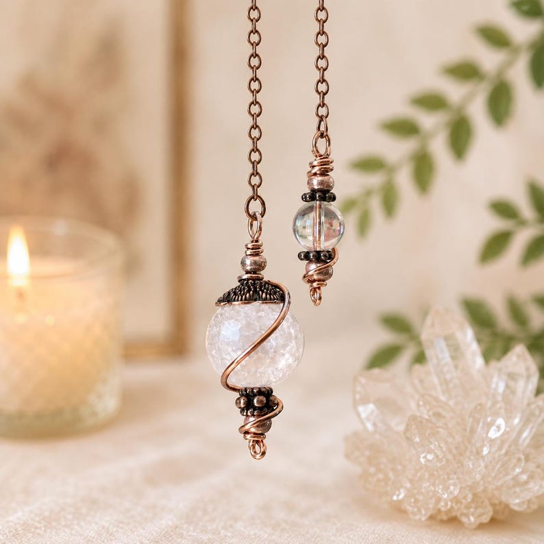 Op de afbeelding: Een koperkleurige metalen ketting hangt aan een grote, bolvormige, gebarsten witte glaskraal omwikkeld met koperdraad, geaccentueerd met donkere metalen kralen. Een kleinere, heldere glaskraal is bevestigd aan een tweede ketting met koperdraad.