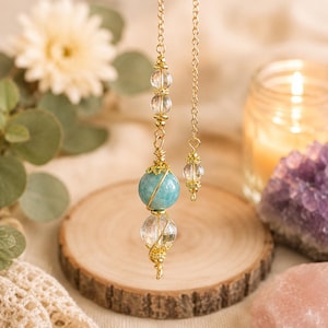 May include: A gold-toned metal chain features a dangling pendant composed of a large, round, opaque teal bead and a smaller, clear, faceted crystal bead, both encased in delicate gold wire wrapping, accented by additional clear crystal beads.