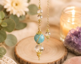 May include: A gold-toned metal chain features a dangling pendant composed of a large, round, opaque teal bead and a smaller, clear, faceted crystal bead, both encased in delicate gold wire wrapping, accented by additional clear crystal beads.