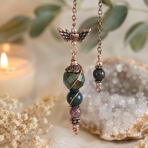 May include: A copper-toned pendulum featuring a winged heart charm at the top, followed by a faceted dark green stone, a smooth dark green bead, a small purple bead, and decorative metal accents, all suspended from a matching copper-toned chain.