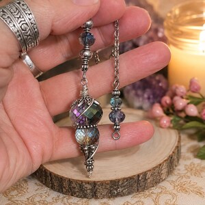 May include: A silver-toned metal pendulum featuring a faceted, iridescent purple and blue glass bead design. The pendulum is suspended from a silver chain adorned with smaller matching glass beads and intricate metal filigree end caps.