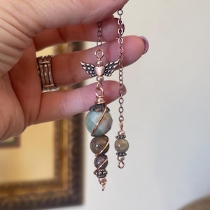 May include: A handcrafted copper pendant featuring a heart with wings, connected to a chain. The pendant has a series of gemstone beads in shades of green, brown, and purple. The design includes a spiral wire detail and a smaller chain with beads.