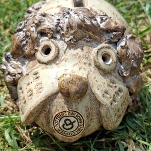 May include: A handcrafted ceramic dog-shaped piggy bank. The bank features a light beige base color with brown accents for the fur and nose. The bank has two eye holes and a slot on top for inserting coins. A circular tag reads "Pottery Made in Hand Cooper".